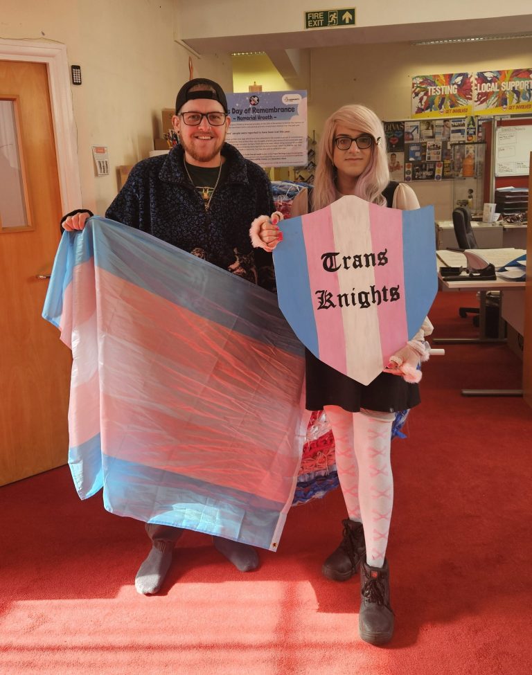 Two people pose with a Trans flag and a shield that says 'Trans Knights' in the Trans flag colours.